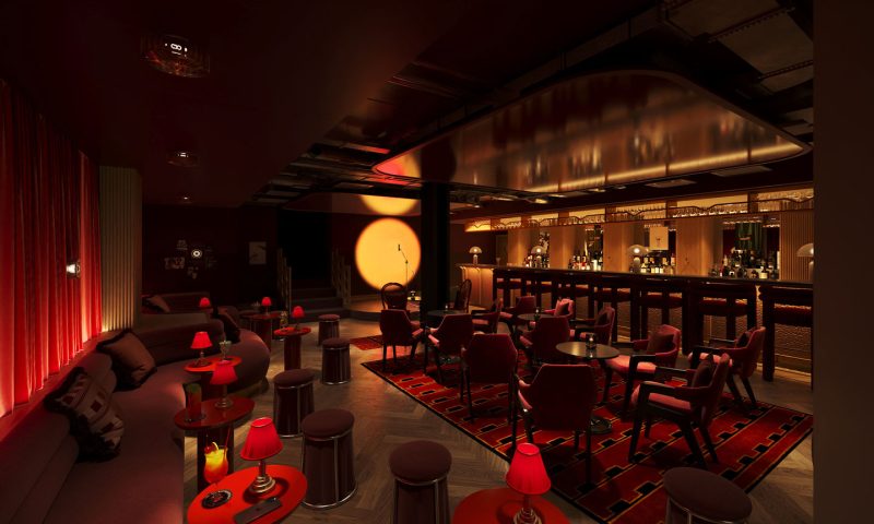 A dimly lit lounge bar in Covent Garden with red chairs, small round tables, red lamps, and a long bar counter. Warm lighting creates a cozy, intimate atmosphere with a spotlight on the back wall and bottles displayed behind the bar.