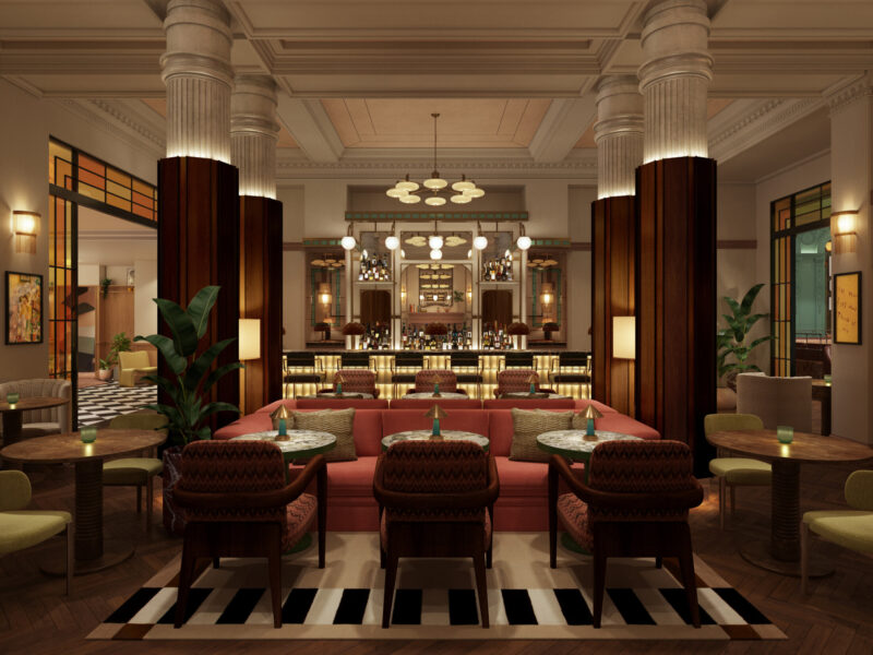 A stylish, upscale lounge at The Webster features plush pink sofas, round tables, patterned chairs, and tall columns. Warm lighting highlights the elegant bar at the back, with plants and modern decor throughout the space.