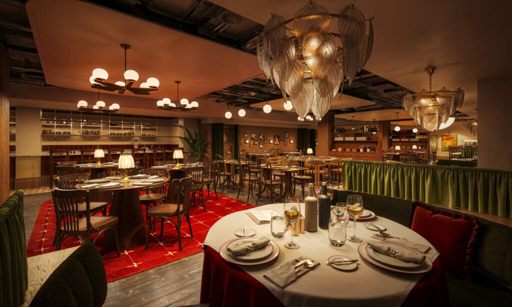 A warmly lit restaurant interior at The Webster features elegant chandeliers, neatly set tables with plates and glasses, wooden chairs, red rug accents, and a cozy, inviting atmosphere with decorative details on the walls.