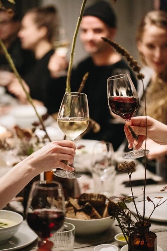 People clink glasses of red and white wine at a dinner table in The Webster, surrounded by food, plates, and dried flowers, with blurred friends in the background enjoying the gathering.