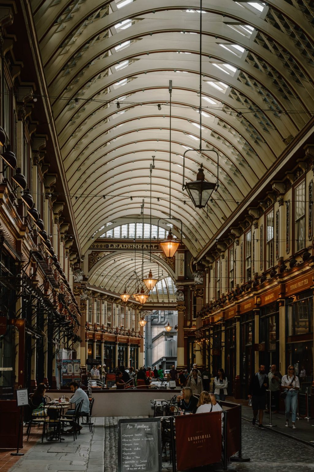A covered Victorian-style shopping arcade with ornate architecture, arched glass ceiling, hanging lamps, and people at café tables. The Webster boutique is among the shops lining both sides of the bustling corridor.