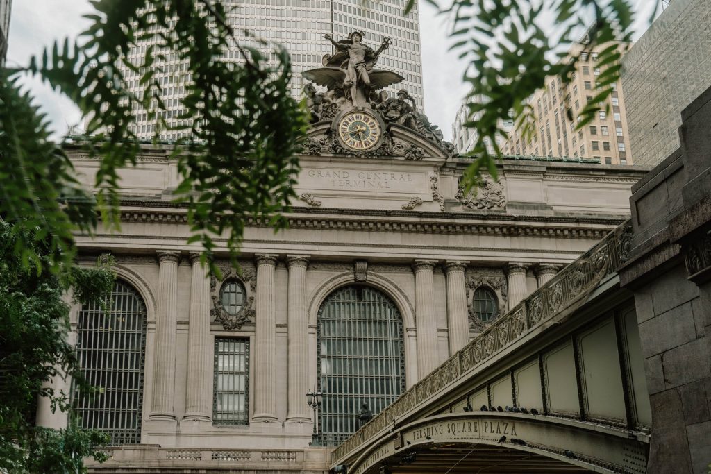 The facade of Grand Central Terminal in New York City, featuring large arched windows, a clock, statues, and nearby buildings like The Webster, is partially framed by green tree branches.