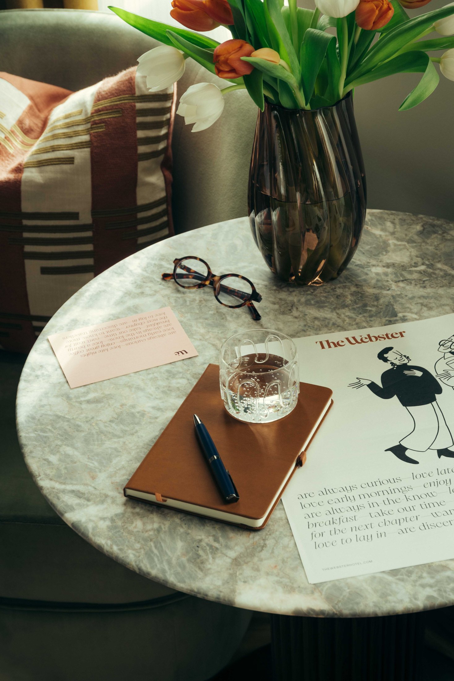 A marble table at The Webster with a brown notebook, pen, glass of water, eyeglasses, a vase of orange and white tulips, a note, and a magazine featuring an illustration of two people titled The Lighter.