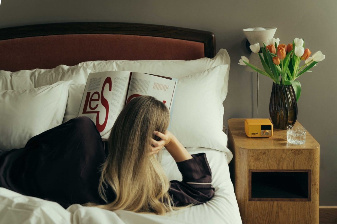 A person with long blonde hair lies on a bed reading a magazine in The Webster. A wooden bedside table holds a vase of tulips, a small yellow radio, and a glass, all set within soft lighting and calm, neutral colors.