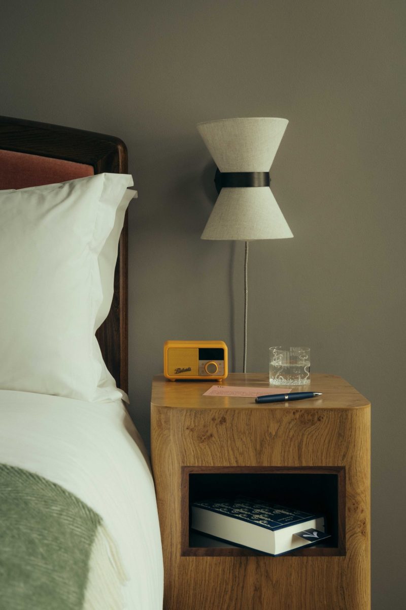 A cozy bedside setup at The Webster features a wooden nightstand holding a yellow vintage radio, a glass of water, notepad, and pen. A book rests on the shelf below, next to a bed with white linens and a modern wall lamp above.