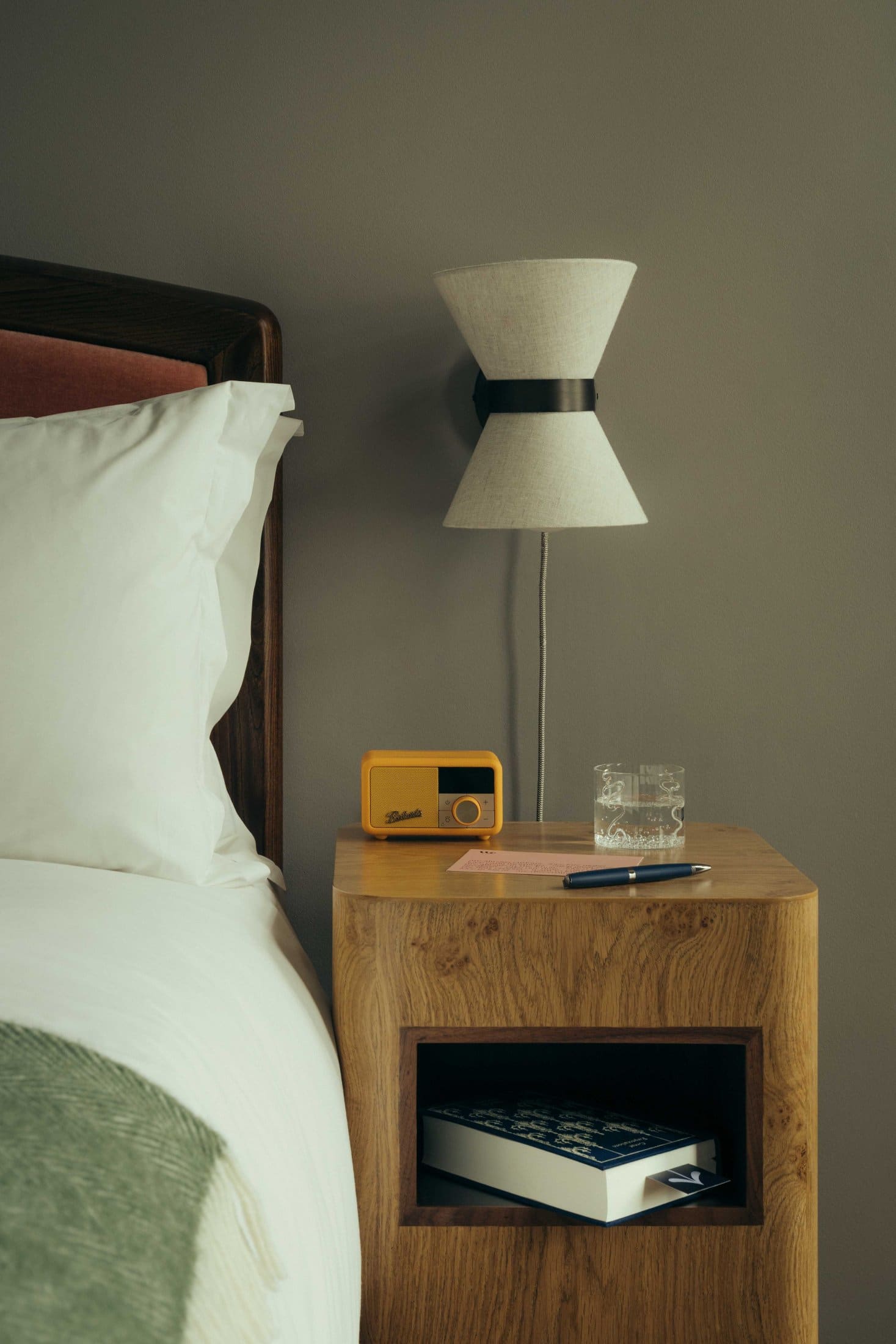 A cozy bedside setup at The Webster features a wooden nightstand holding a yellow vintage radio, a glass of water, notepad, and pen. A book rests on the shelf below, next to a bed with white linens and a modern wall lamp above.
