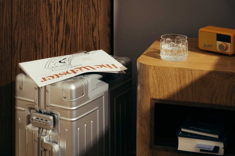 A silver suitcase with a magazine on top stands beside a wooden nightstand from The Webster, holding a glass of ice water, an orange radio, and several books inside an open shelf. Warm light creates soft shadows across the scene.