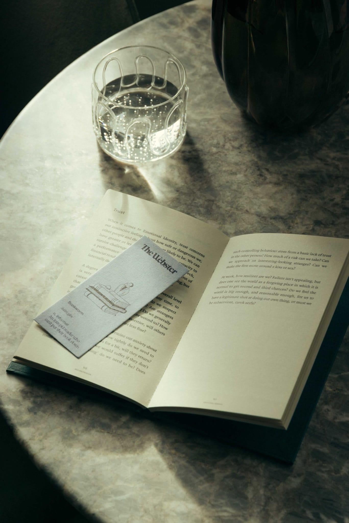 An open book from The Webster with a bookmark and a glass of water sit on a round marble table in soft, natural light.