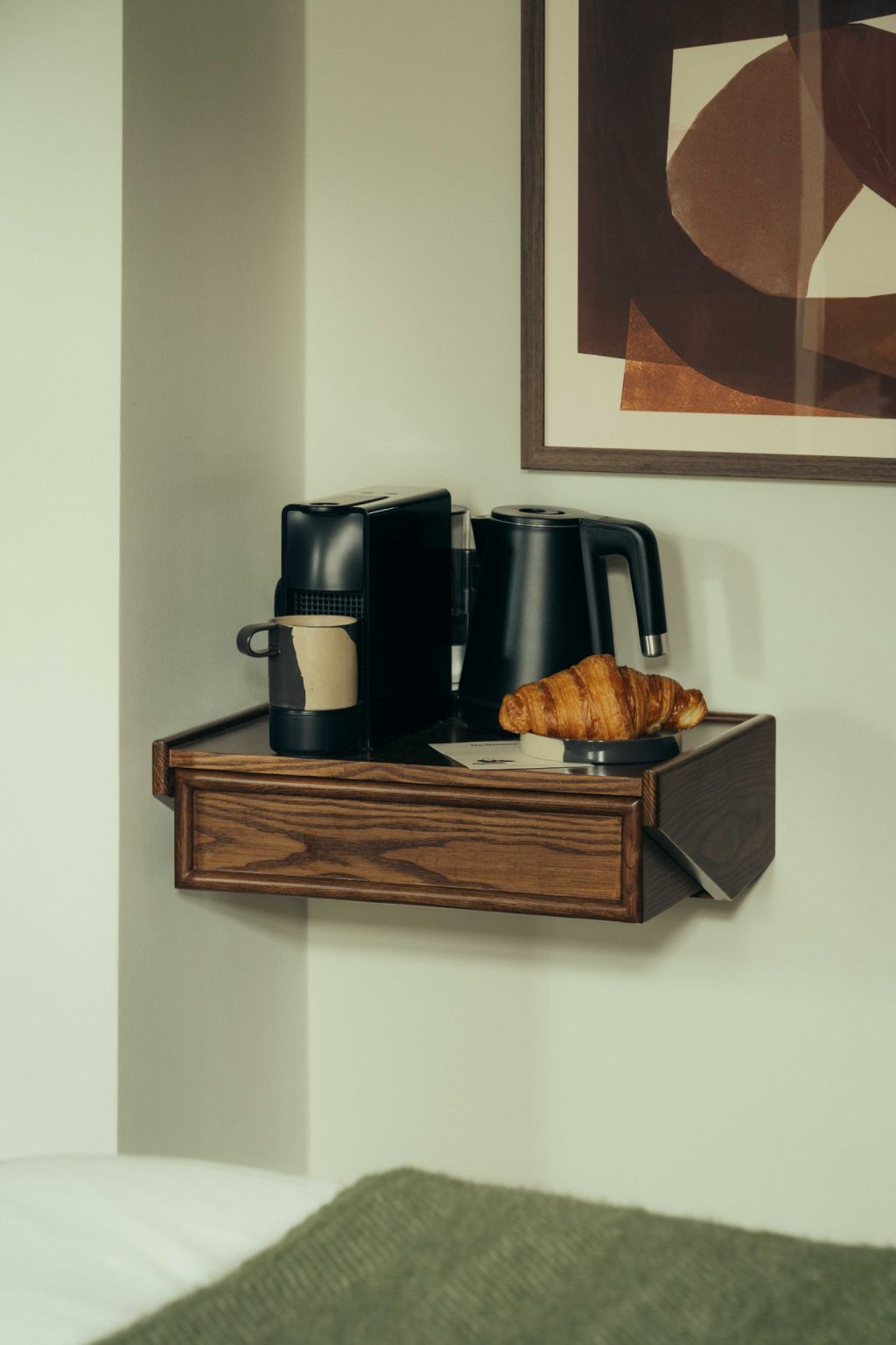 A small wooden shelf in a corner at The Webster holds a coffee machine, a black electric kettle, a cup with a sleeve, a croissant on a plate, and a postcard. A modern abstract artwork hangs above the shelf.