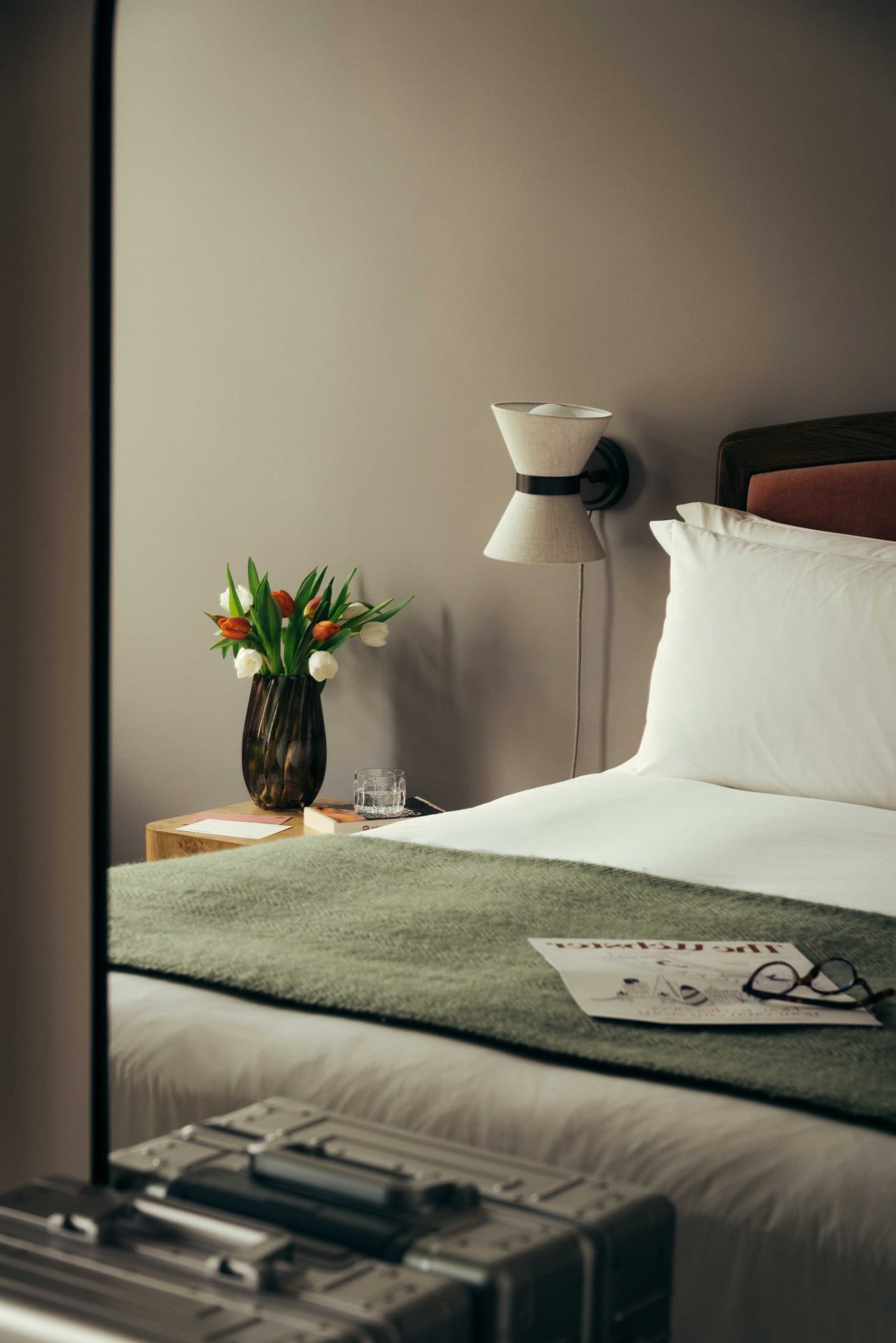 A neatly made bed with white sheets and a green blanket at The Webster, next to a nightstand holding a vase of tulips, a glass of water, and a wall-mounted lamp; a suitcase sits at the foot of the bed.