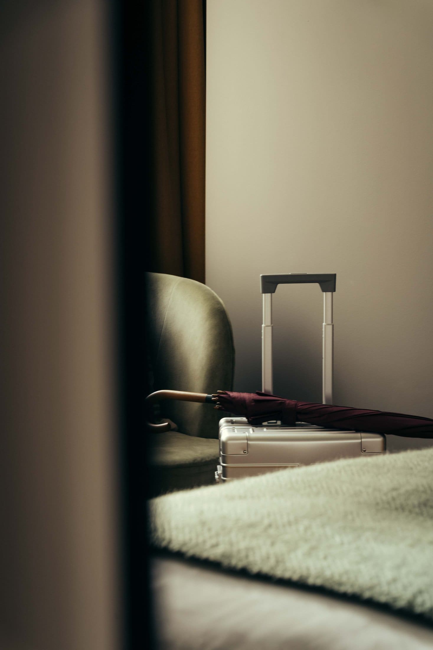 A soft-focused image of a room at The Webster shows a green chair, a closed red umbrella, a metallic suitcase, and the extended handle of rolling luggage next to a green bedspread, with warm light on beige walls.