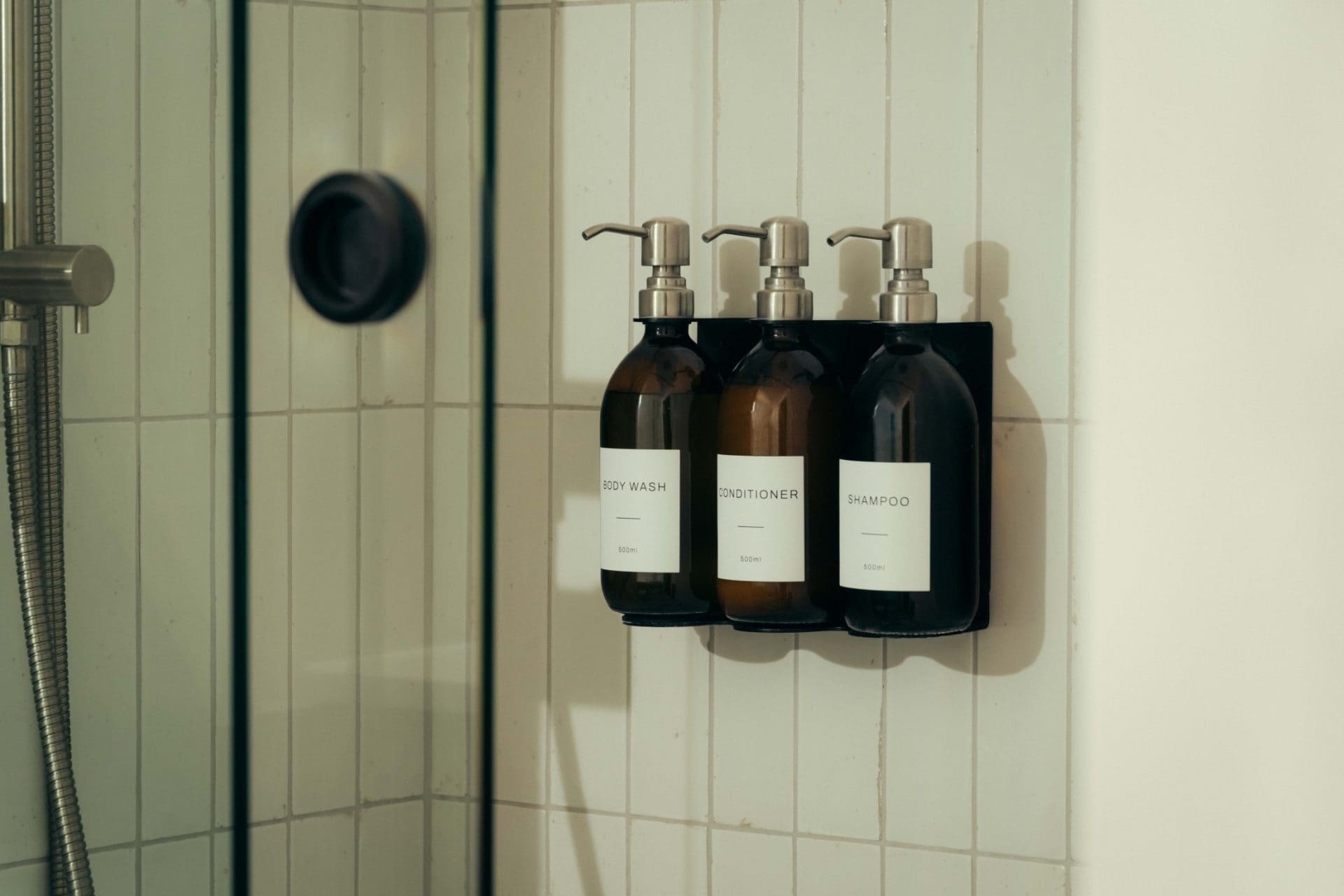 Three pump bottles labeled body wash, conditioner, and shampoo are mounted on a black wall rack inside a tiled shower at The Webster. A shower control knob and part of a handheld showerhead are visible nearby.