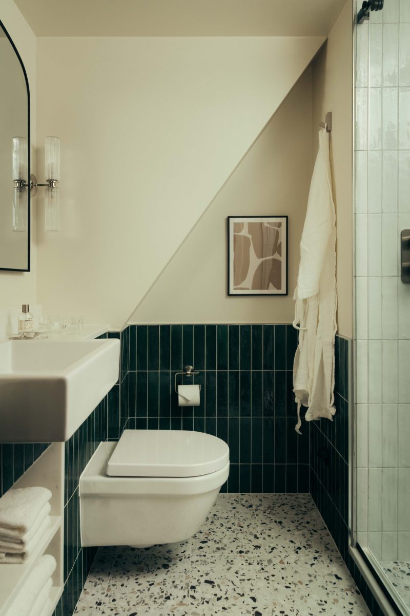 Modern bathroom at The Webster with green tiled walls, a floating white sink, wall-mounted toilet, terrazzo floor, glass shower, abstract wall art, and a white towel hanging by the shower under a slanted ceiling.