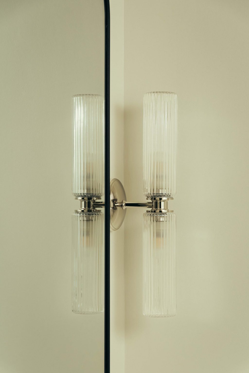 A modern wall sconce, The Webster, features two ribbed glass shades and is mounted on a light-colored wall beside a mirror, creating a symmetrical reflection of the fixture.