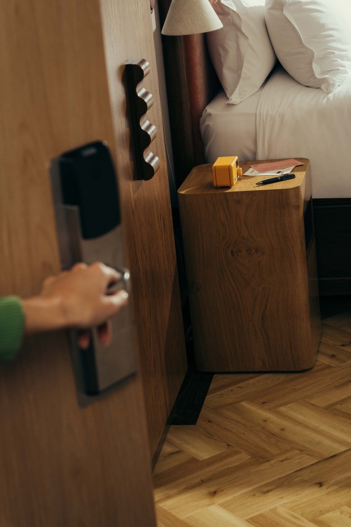 A hand opens a wooden door to a cozy bedroom at The Webster, with a neatly made bed, a wooden nightstand holding a yellow camera, sunglasses, and a phone. The floor displays a light wood herringbone pattern.