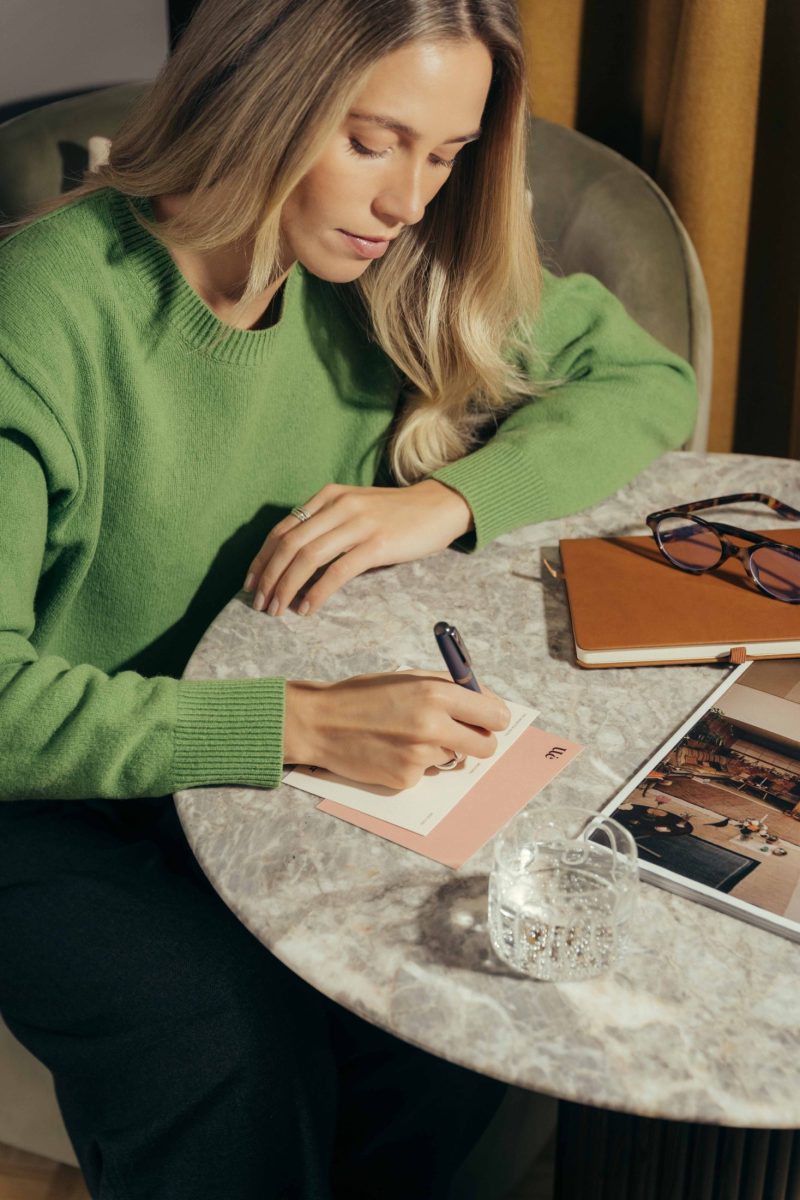 A woman in a green sweater sits at a marble table at The Webster, writing on a notecard. Nearby are a notebook, eyeglasses, a magazine, and a glass of water.