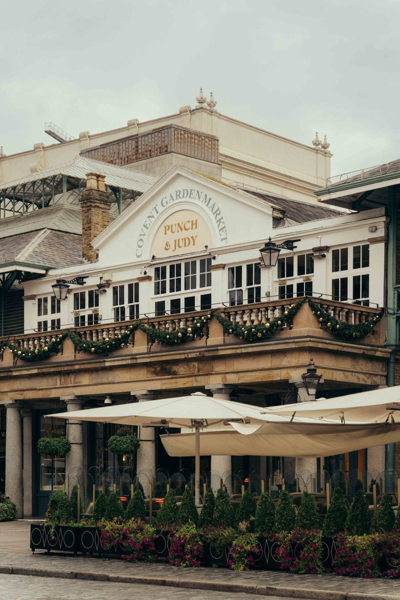 Covent Garden Market’s Punch & Judy pub exterior, much like The Webster, features decorated garlands, classic lanterns, and white awnings over outdoor seating, with potted plants and flowers lining the charming patio.