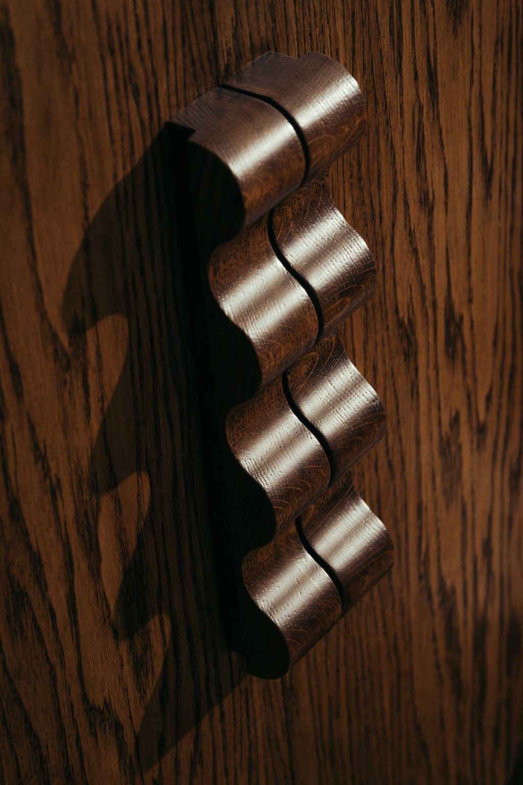A close-up of The Webster, a wavy, sculpted wooden door handle attached to a wood-grain door, casting an elegant shadow on the surface.