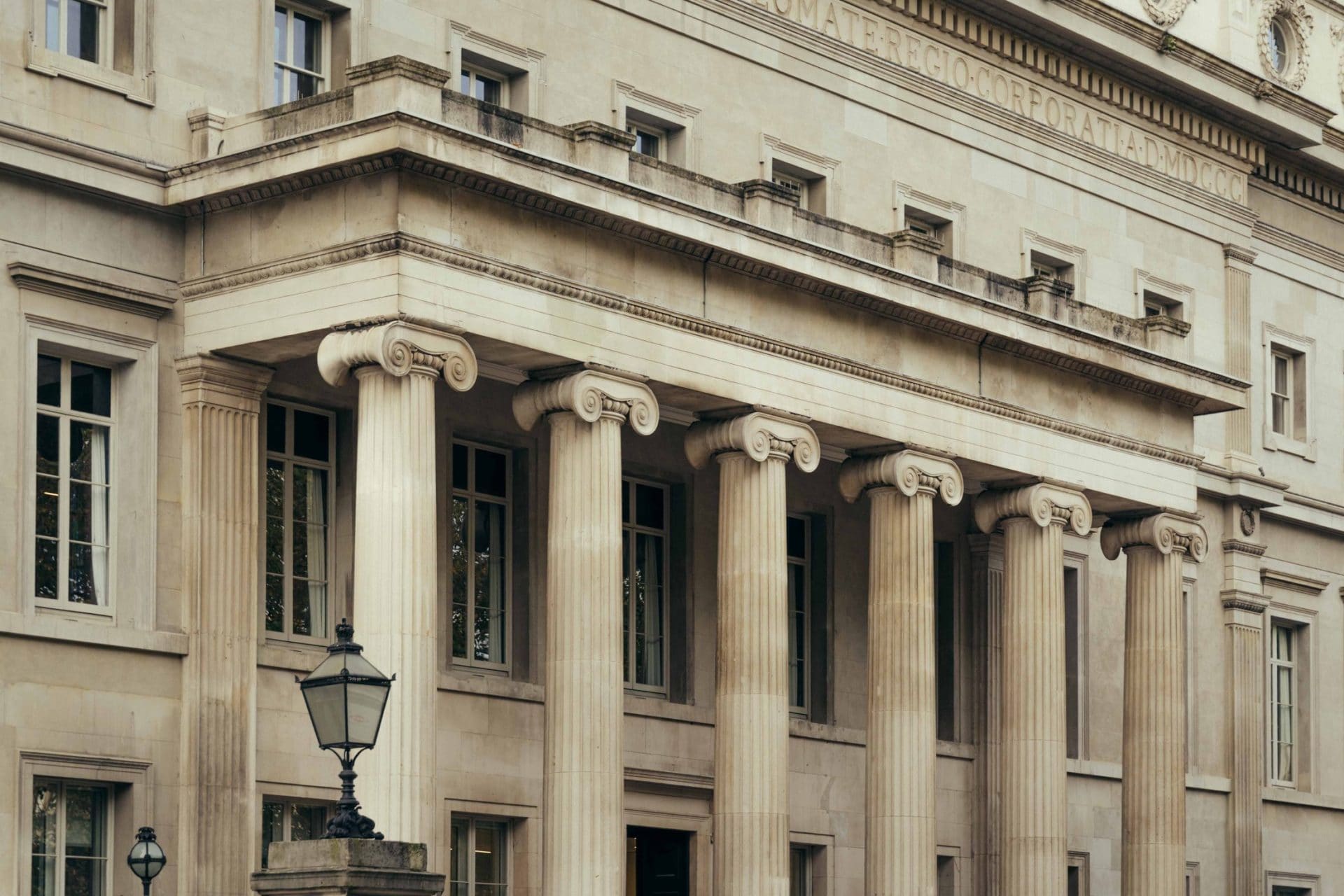 The Webster features a neoclassical building facade with tall, fluted columns topped by Ionic capitals, rectangular windows, and an old-fashioned street lamp in front. The building is made of light-colored stone.