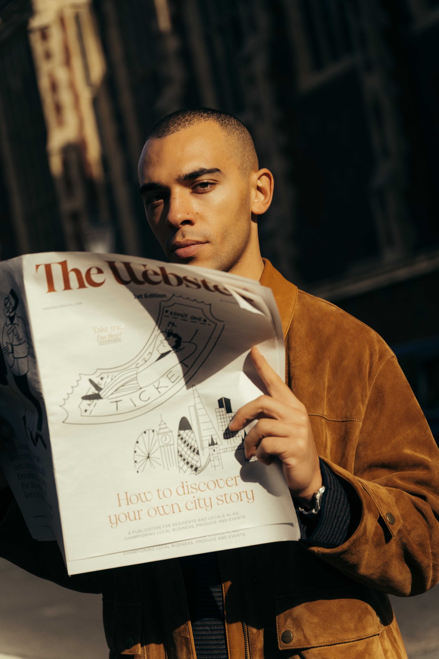 A man in a brown suede jacket stands outdoors, holding and reading The Webster newspaper. Sunlight highlights his face as he looks up from the paper, with a blurred building in the background.