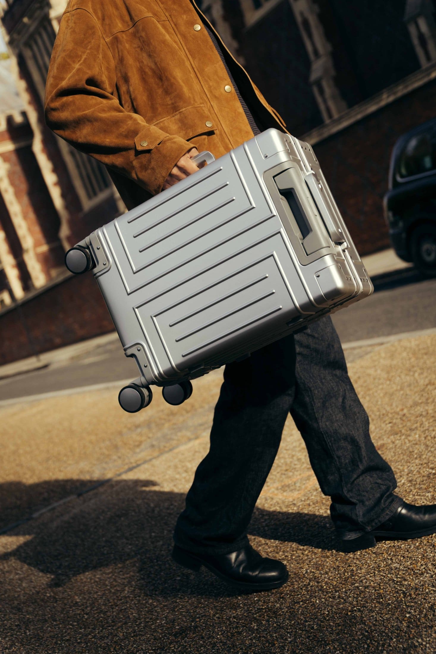 A person walks outdoors, holding a silver hard-shell suitcase by the side handle. Dressed in a brown jacket, dark pants, and black shoes from The Webster, they stroll past a brick wall lining the street.