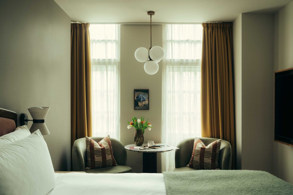 A cozy bedroom at The Webster with two large windows, mustard curtains, a bed, two armchairs with patterned cushions, a small round table topped with tulips, and globe pendant lights overhead.