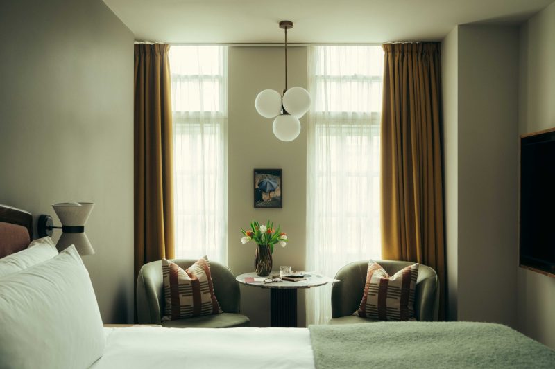 A cozy bedroom at The Webster with two large windows, mustard curtains, a bed, two armchairs with patterned cushions, a small round table topped with tulips, and globe pendant lights overhead.