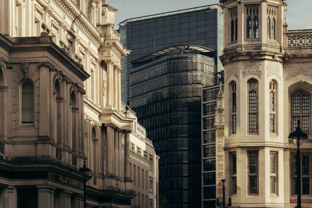 Historic stone buildings with ornate facades frame a view of The Webster, a modern glass and steel skyscraper, rising in the background under a cloudy sky.