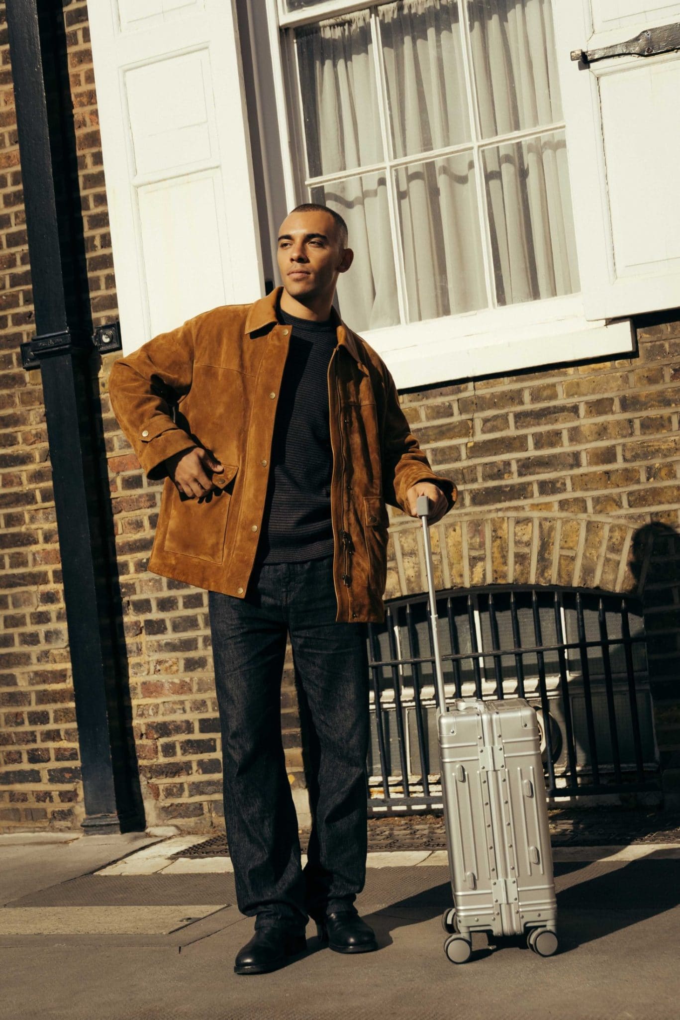 A person stands outside The Webster by a brick building, wearing a brown jacket, black shirt, and jeans, with one hand resting on the handle of a silver rolling suitcase.