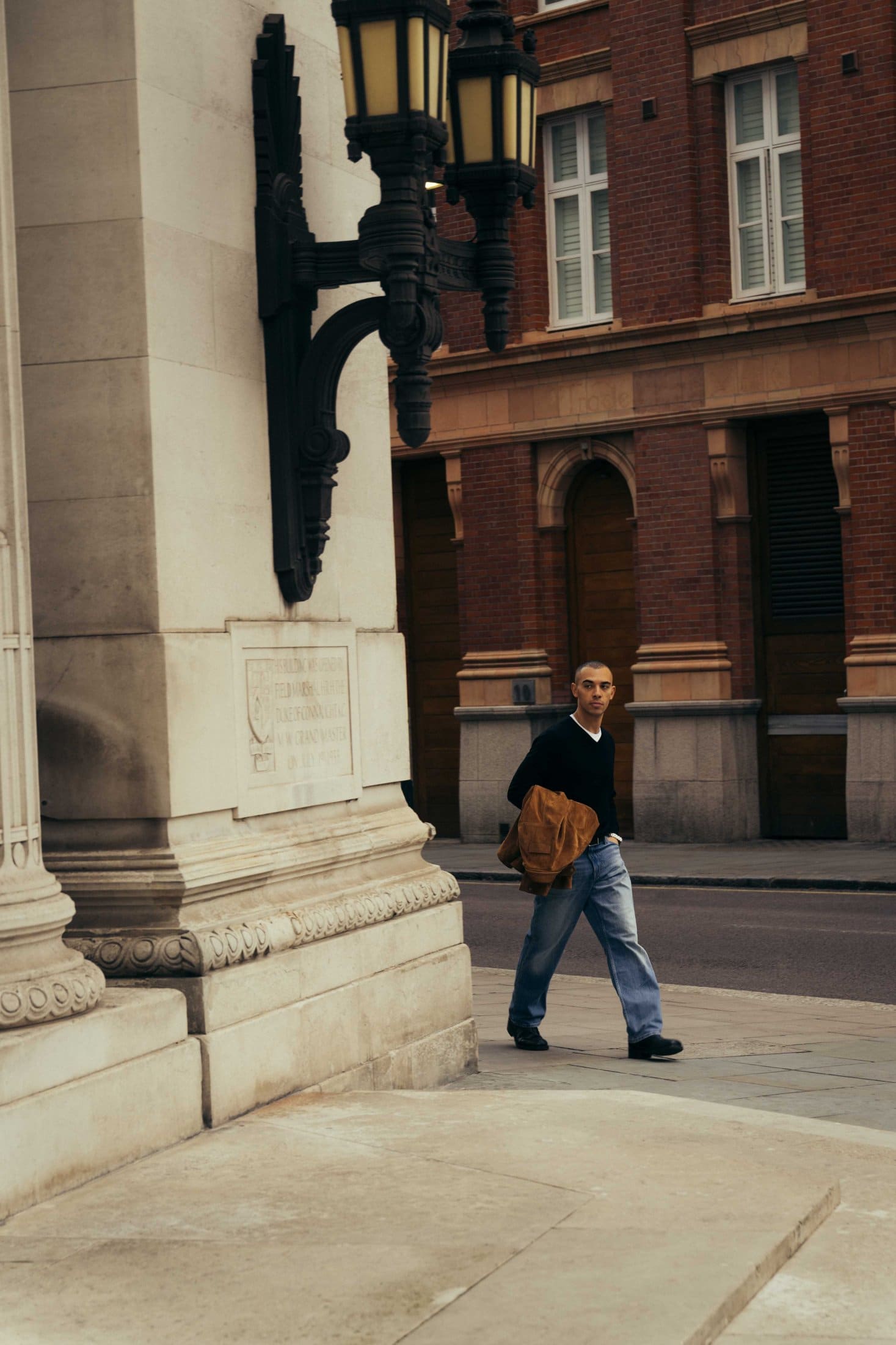 A person wearing a black sweater and blue jeans walks on a city sidewalk near The Webster, holding a brown jacket. The scene features historic stone and brick buildings and ornate street lamps.
