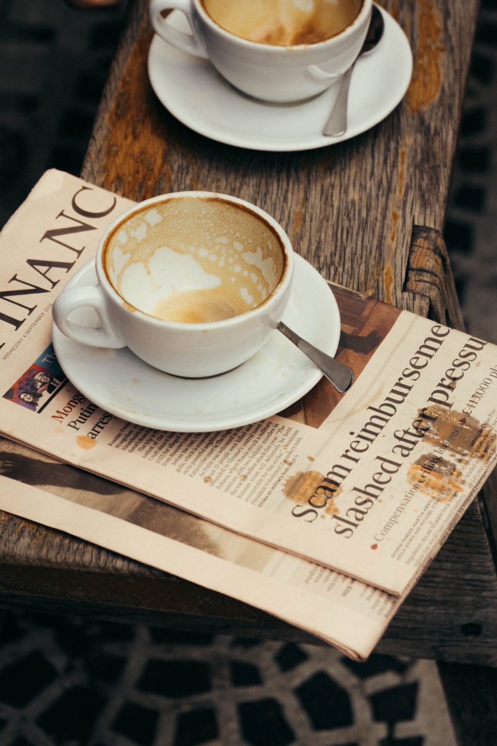A nearly empty cappuccino cup sits on a saucer with a spoon atop financial newspapers at The Webster, resting on a rustic wooden table. Another cup is partially visible in the background.