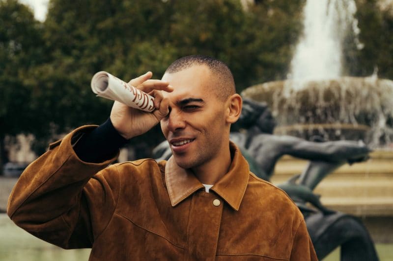 A man in a brown suede jacket holds a rolled-up paper to his eye like a telescope and smiles, standing in front of The Webster fountain with a statue and trees in the background.
