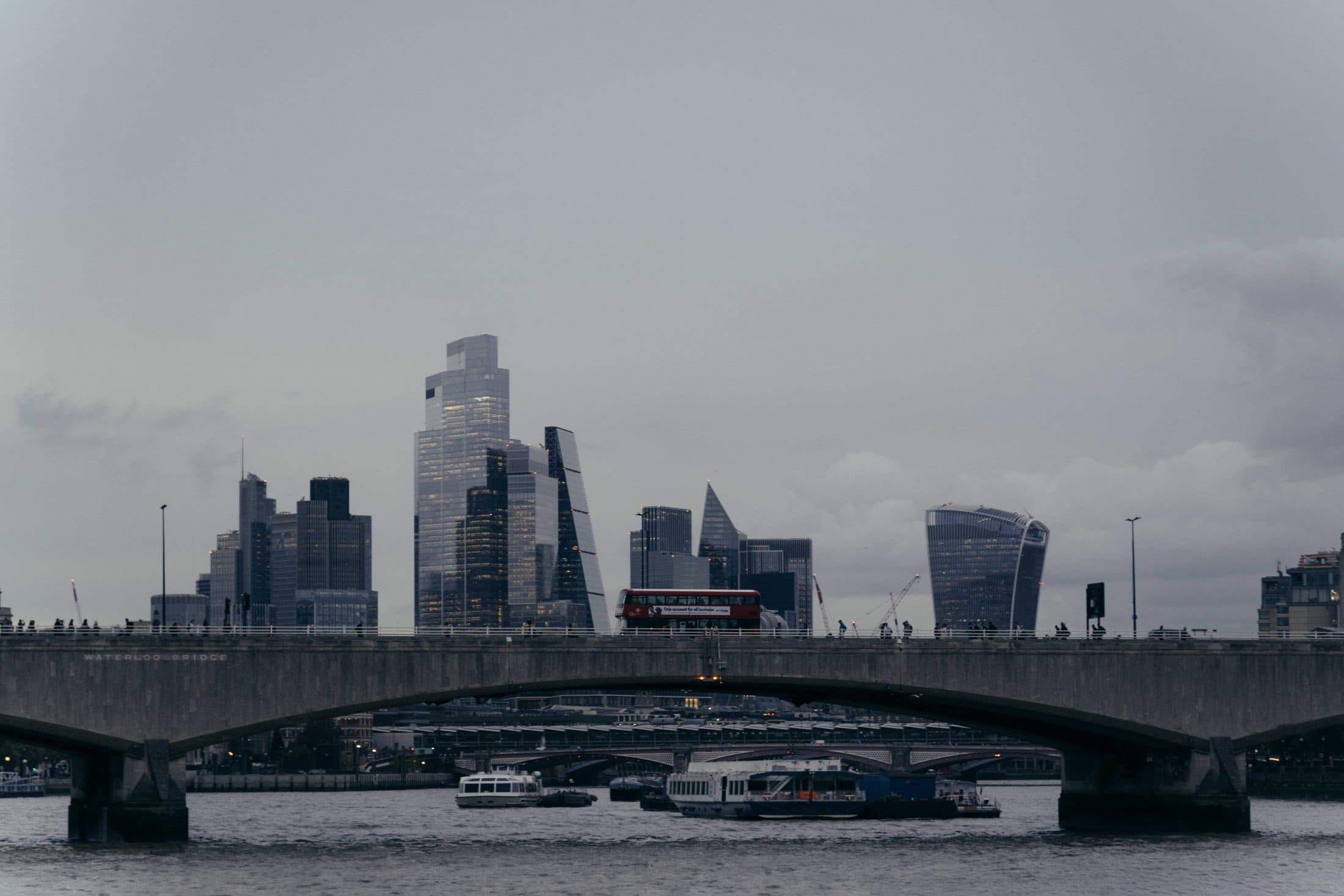 A cityscape of London at dusk, featuring modern skyscrapers in the background, a red double-decker bus crossing The Webster Bridge, and boats passing underneath on the River Thames.