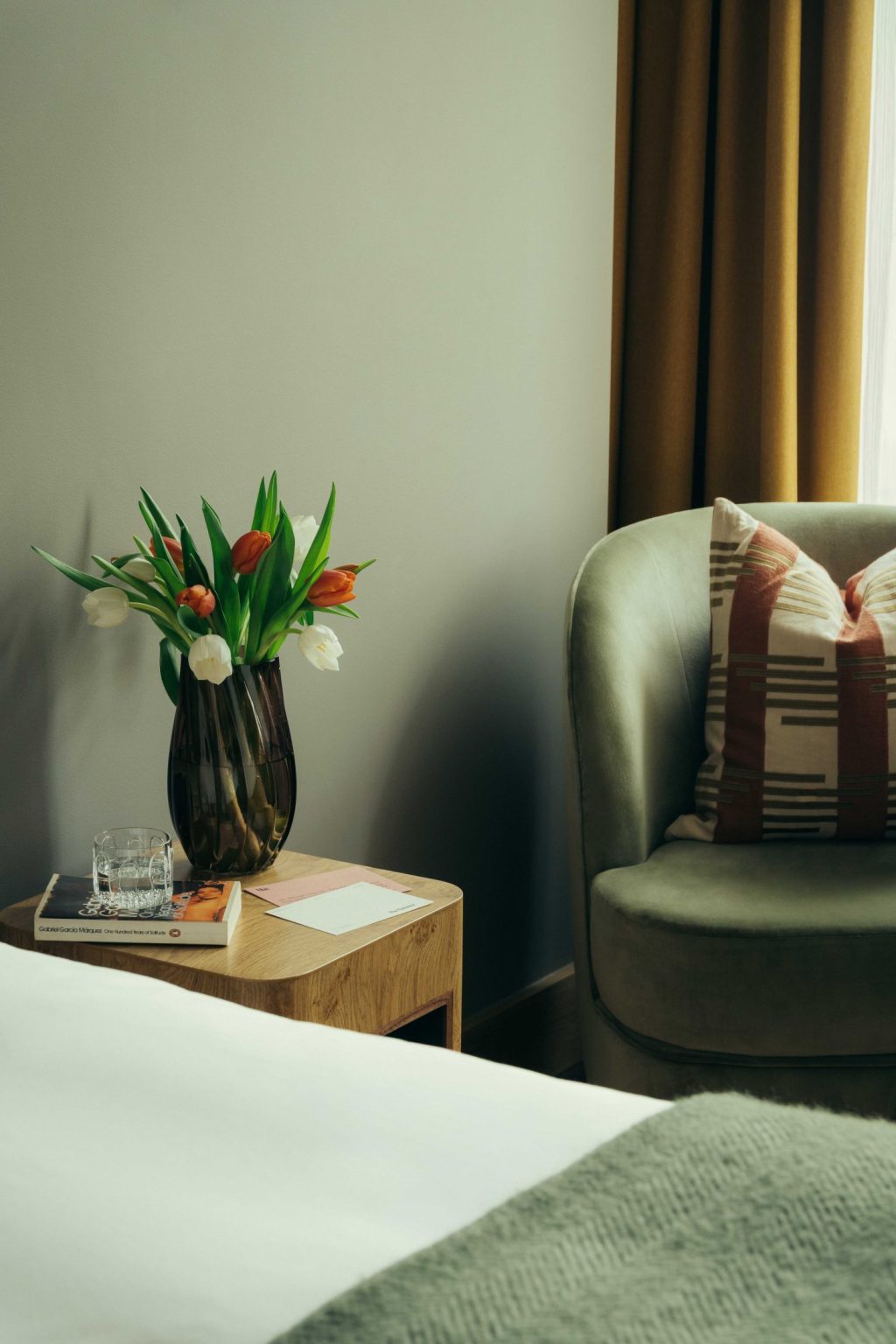 A cozy living space at The Webster features a green armchair, patterned cushion, side table with a vase of tulips, a glass of water, and books beside a bed draped in a green blanket.