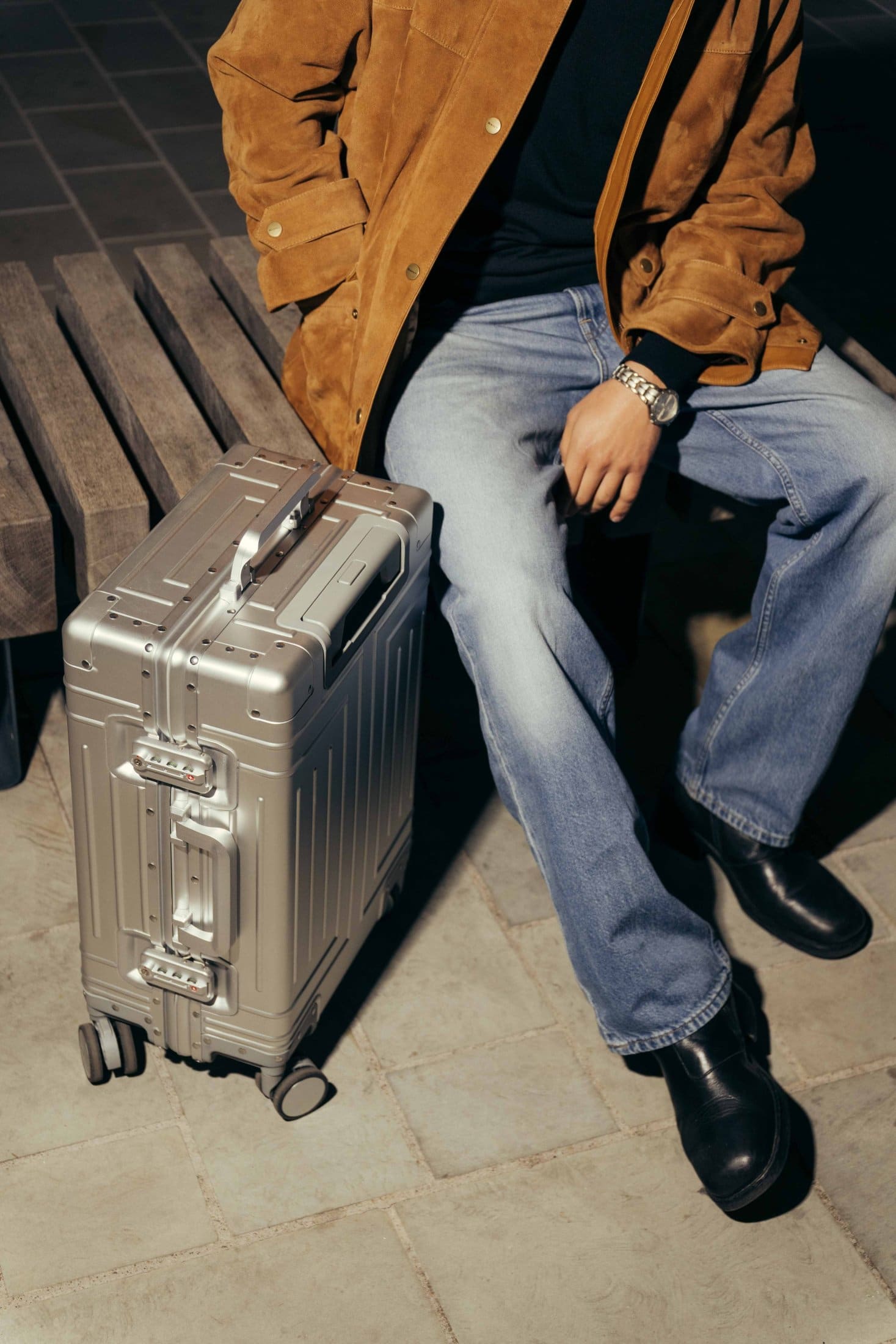 A person wearing a brown jacket, jeans, and black boots sits on a bench next to a silver rolling suitcase on a tiled floor at The Webster. Their face is not visible.