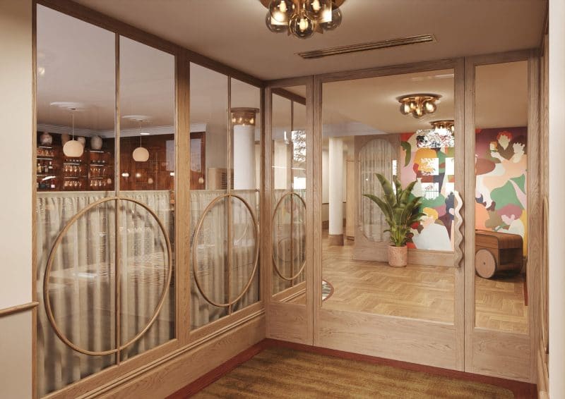A hallway at The Webster with wood and glass walls featuring circular designs opens into a warmly lit room with wooden floors, modern chandeliers, a large plant, and a colorful abstract mural.