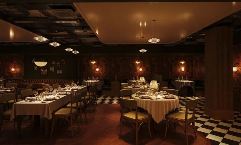 A dimly lit, elegant restaurant at The Webster features round tables draped in white cloths, set for dining. Warm lighting, checkered and wooden floors, and ornate wallpaper create a cozy, vintage atmosphere.