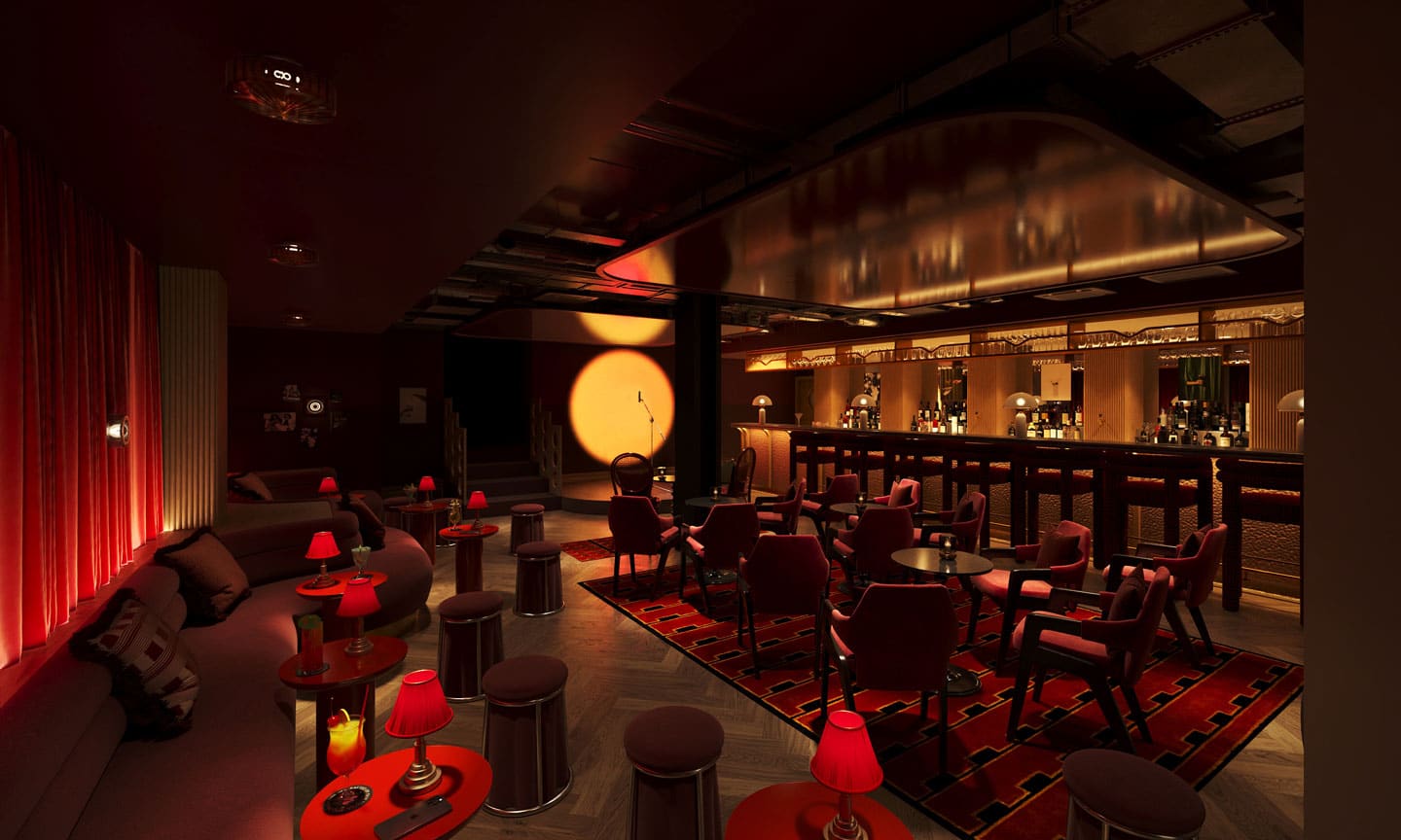 A dimly lit lounge bar at The Webster, featuring red lighting, small round tables, red chairs, and red lamps. A spotlight shines on the wall near the bar area, creating a cozy, intimate atmosphere with shelves stocked with bottles and glasses.