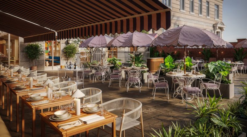 A stylish outdoor restaurant patio at The Webster, featuring striped umbrellas and awnings, elegant tables set with plates and glasses, lush green plants, and a building in the background on a sunny day.
